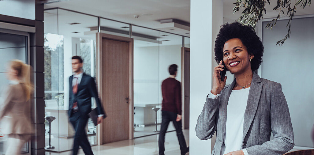 black business woman on her cell phone in an office lobby with four other office workers passing by in the background.