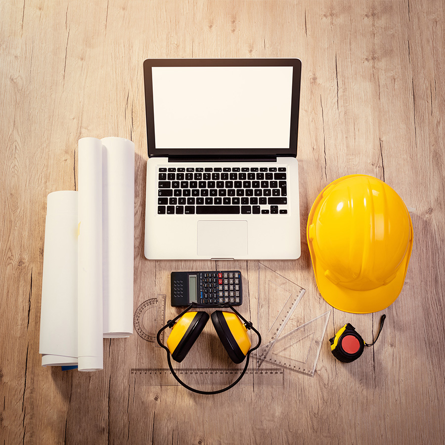 composed overhead photo of a laptop surrounded by a hard hat, ear protection, calculator, tape measure, ruler, square, and rolled up paper.