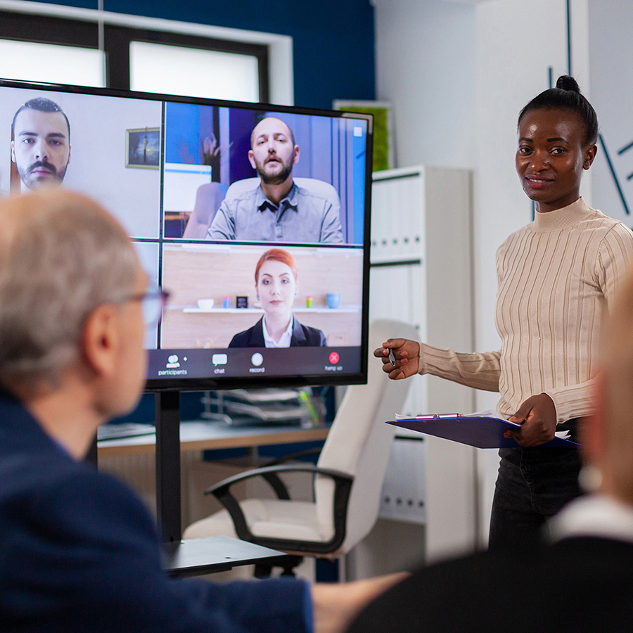 woman facilitating a training session with an older man and another partially obscured colleage in the room. she is standing in front of a video display showing two male and one female remote participants.