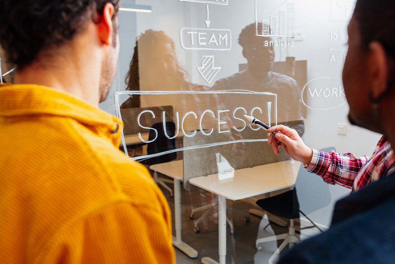 several colleagues of different races and sexes looking at a clear drawing board with the word "success!" written in white marker.